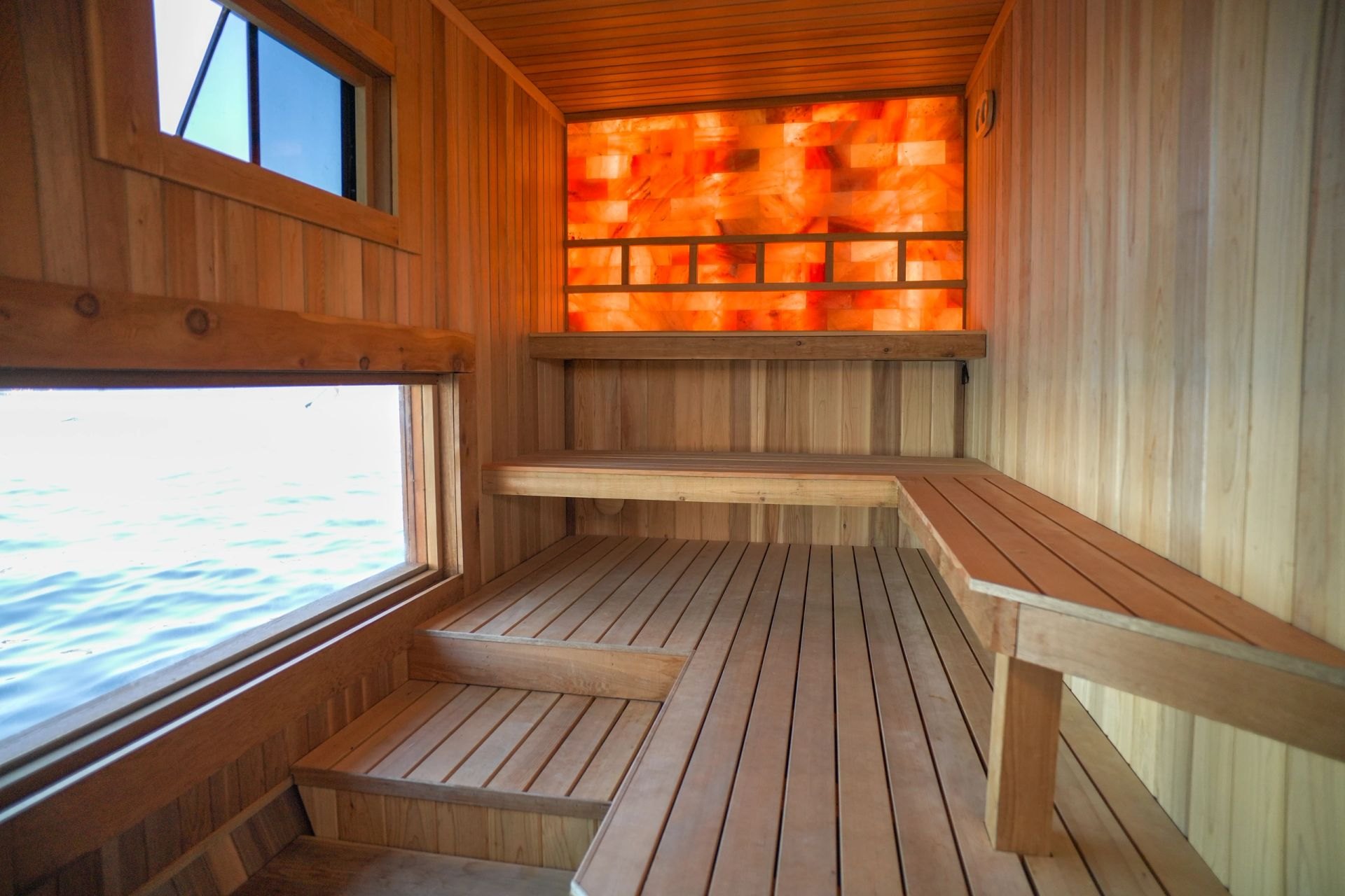 Cedar sauna benches beneath the glowing Himalayan salt wall, with a window over the water